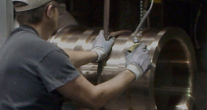 Machinist uses a tool while working on a large bronze cylinder mounted in a machine.