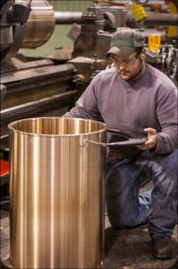 Machinist kneels beside a tall machined bronze cylinder, checking a matching cover or ring.