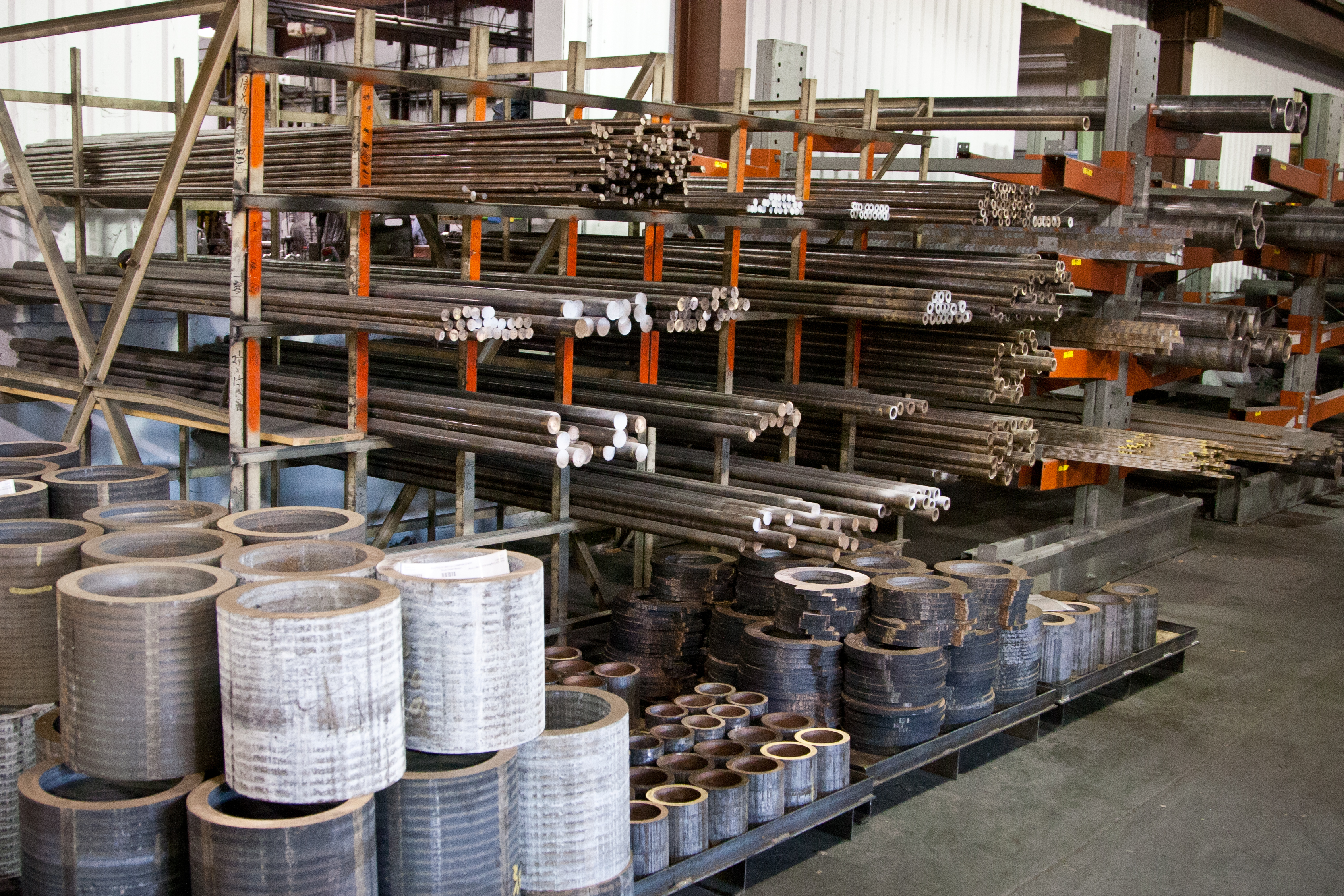 Warehouse inventory with metal bars and tubes on racks and large ring sections staged on the floor.
