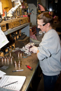 Employee finishes small machined metal parts at a workbench with measuring tools and shop supplies.